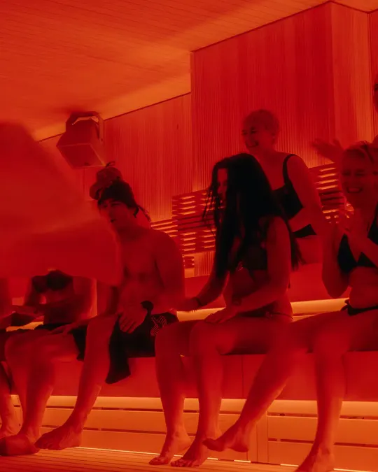 A crowd in swimsuits on the benches of a red-lit sauna at Othership, one person fanning the heat with a towel